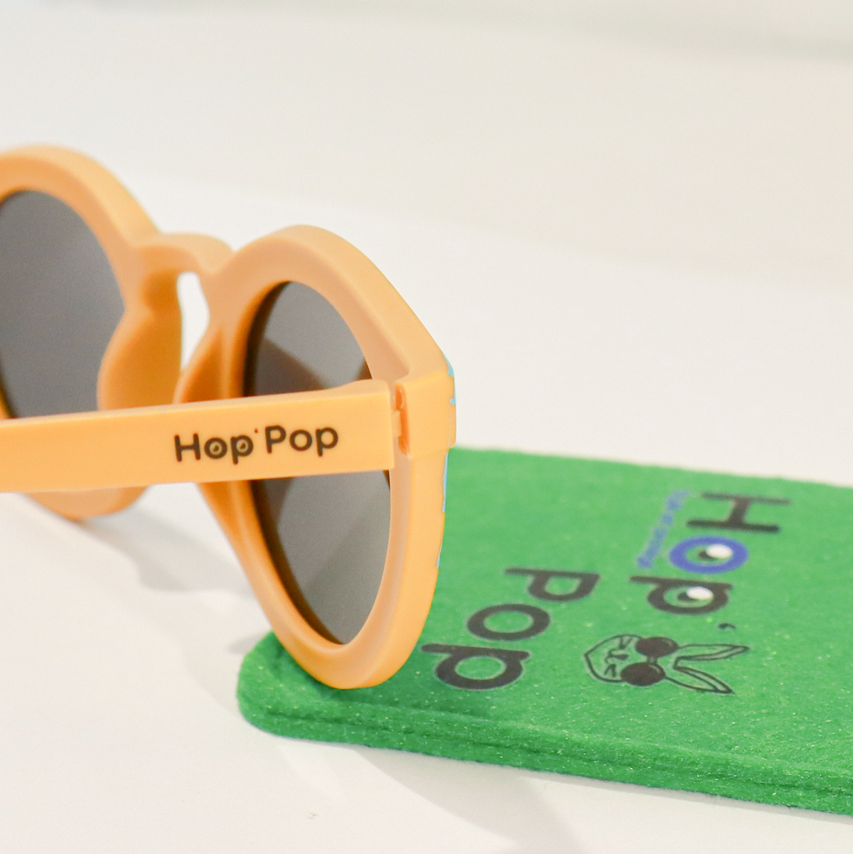 Gros plan sur une paire de lunettes de soleil orange de la marque Hop'Pop. Le logo de la marque est visible en noir sur la branche latérale. Les verres teintés, probablement polarisés, suggèrent une protection solaire élevée. À l’arrière-plan flou, on distingue une pochette de rangement verte avec des illustrations ludiques, assortie à l’univers enfantin ou familial de la marque.