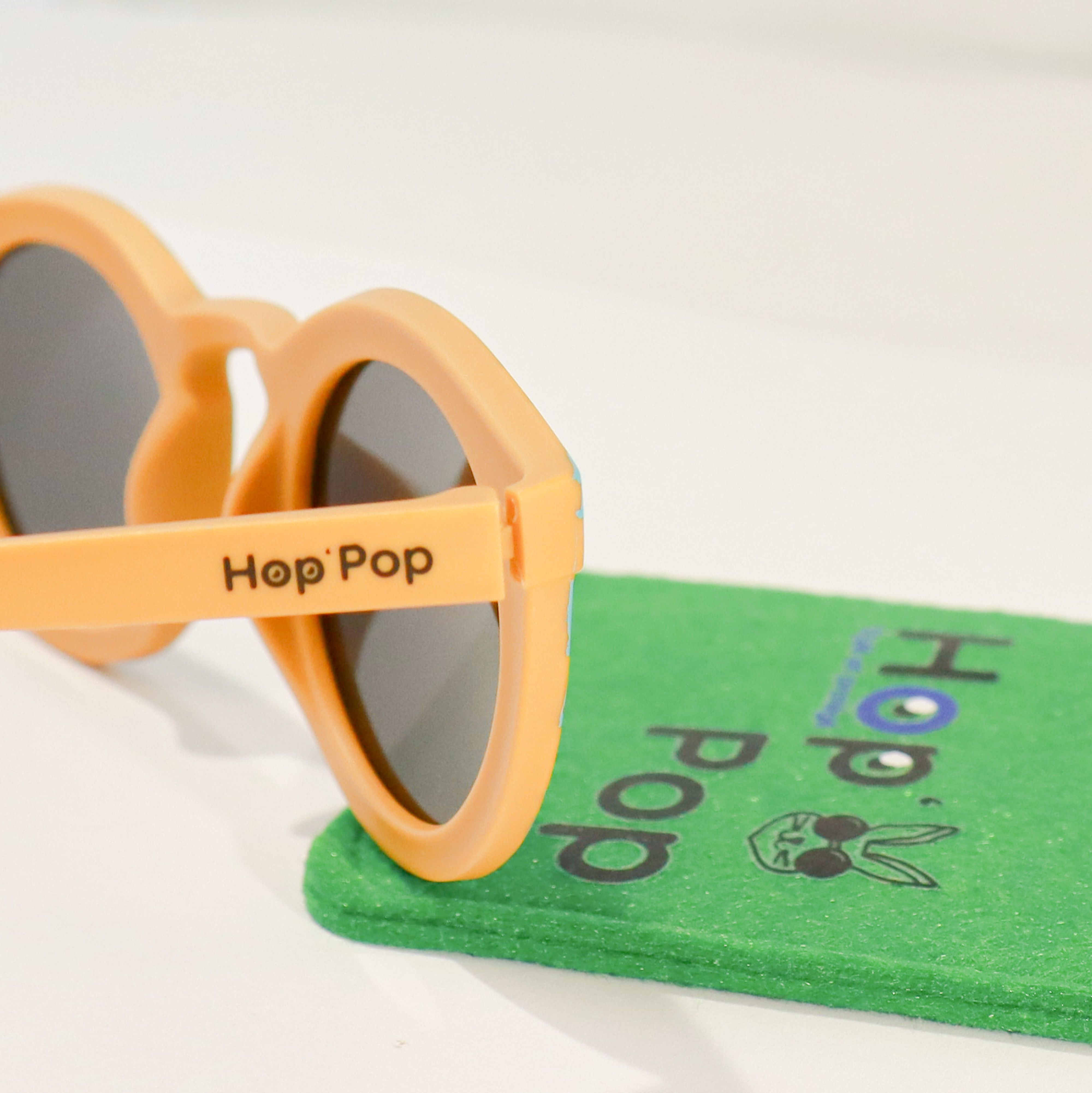 Gros plan sur une paire de lunettes de soleil orange de la marque Hop'Pop. Le logo de la marque est visible en noir sur la branche latérale. Les verres teintés, probablement polarisés, suggèrent une protection solaire élevée. À l’arrière-plan flou, on distingue une pochette de rangement verte avec des illustrations ludiques, assortie à l’univers enfantin ou familial de la marque.